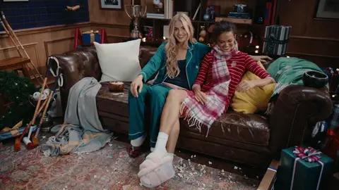 Two women sit together on a brown leather sofa, one in teal loungewear and the other in red stripes with a pink plaid scarf.