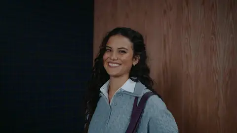 A smiling woman with dark curly hair wears a gray cable-knit polo sweater and a burgundy shoulder strap against blue plaid and wood panels.