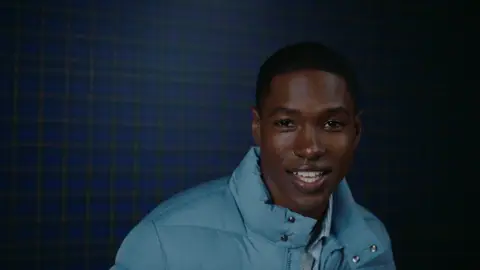 A smiling man wearing a light blue puffer jacket leans toward the camera against a dark blue plaid background.