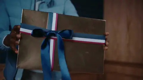 Hands hold a gold wrapped gift with navy ribbon and red-white-blue trim in front of a light blue puffer jacket.