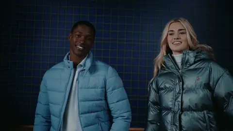 A man in a light blue puffer jacket and a woman in a metallic green puffer jacket stand together against blue plaid panels.