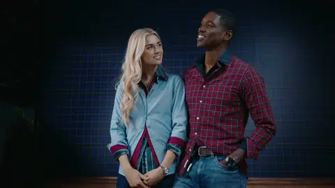 A woman in a light blue button-down shirt looks toward a smiling man in a red plaid shirt against a blue plaid background.