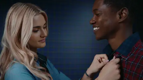 A woman in a light blue shirt adjusts the collar of a smiling man wearing a red plaid shirt.