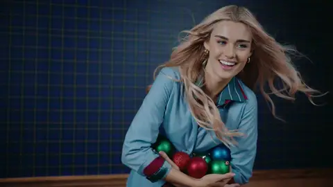 A woman in a light blue shirt holds red, green and blue holiday ornaments as her long blonde hair moves to one side.