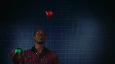 A man in a red plaid shirt looks upward while juggling red and green holiday ornaments against a blue plaid background.