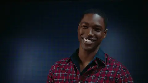 A smiling man in a red plaid button-down shirt faces the camera in a close portrait against dark blue plaid panels.