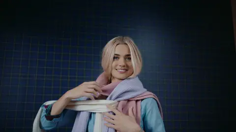 A smiling woman in a light blue shirt holds layered pink, lavender and cream scarves around her shoulders against blue plaid panels.