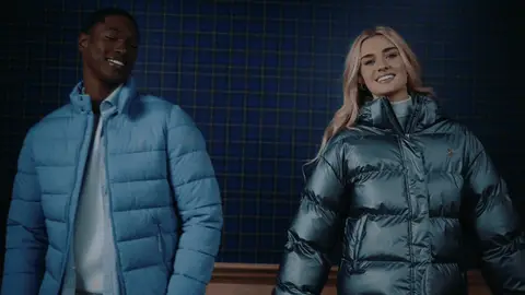 Two adults turn and smile in light blue and metallic green puffer jackets against a blue plaid background.