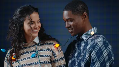 A woman in a patterned cream sweater smiles at a man in a navy plaid-knit sweater, with colorful holiday lights draped across them.