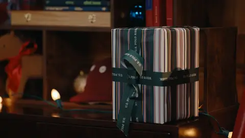 A striped gift box tied with a green ribbon sits beside a wooden bookcase and a glowing holiday light.