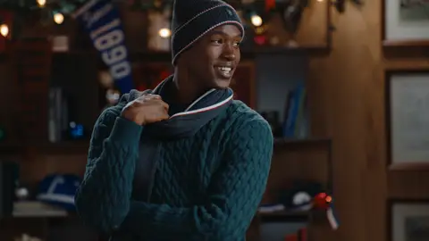 A smiling man wears a deep green cable-knit sweater, dark beanie and striped scarf in front of a bookcase with holiday lights.