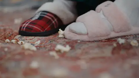 Red plaid slippers and pale pink fuzzy slides rest on a patterned rug scattered with popcorn.