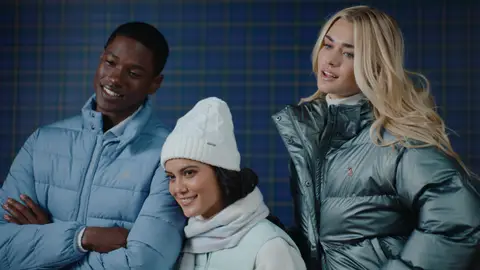 Three adults pose together in a pale blue puffer jacket, mint vest with a white beanie, and metallic green puffer jacket against a plaid wall.
