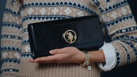A woman holds a black embossed clutch with a round gold rider emblem against a cream patterned sweater, with a gold watch at her wrist.