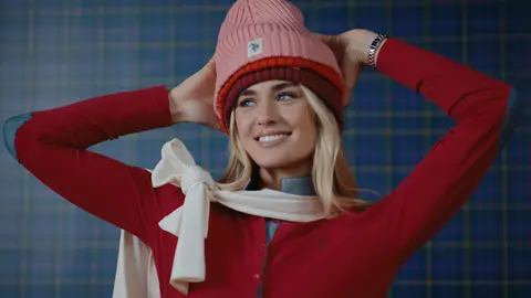 A smiling woman holds stacked pink, orange and burgundy knit beanies above a red top and a cream scarf tied at her shoulder.