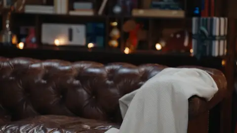 A cream throw with muted green stripes hangs over the arm of an empty tufted brown leather sofa in warm interior light.