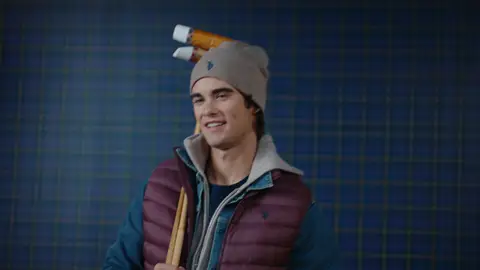 A man in a taupe beanie and burgundy quilted vest over denim and gray layers carries two polo mallets at his shoulder.