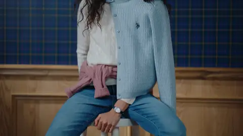 A seated woman wears pale blue and white knit layers, a pink sweater tied at her waist and blue jeans, with a wristwatch above her resting hands.