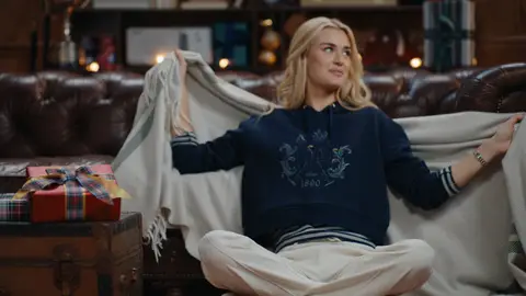 A woman in a navy hoodie and white trousers sits cross-legged on a patterned rug, holding a cream throw open behind her.