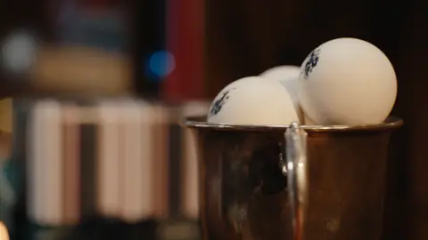 Two white balls with blue rider logos sit in a silver trophy cup against a softly lit wooden bookcase.