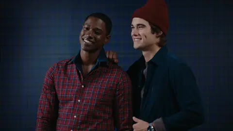 Two men smile together against a blue checked wall, one in a red plaid shirt and the other in navy layers with a burgundy beanie.