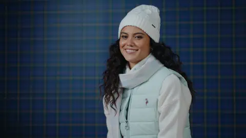 A smiling woman wears a mint quilted vest over a cream top, a white cable-knit beanie and a pale checked scarf against a blue plaid wall.