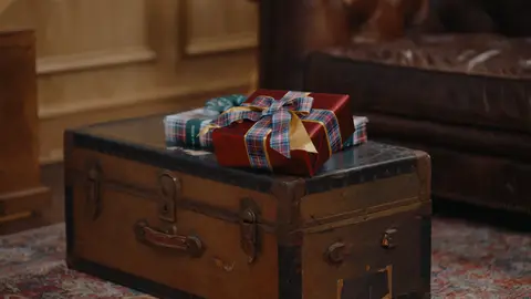 Plaid-wrapped gifts rest on a weathered wooden trunk beside a brown leather sofa and a patterned rug.