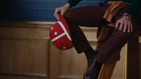 A seated man holds a red polo helmet beside burgundy trousers and polished dark boots on a wooden seat.
