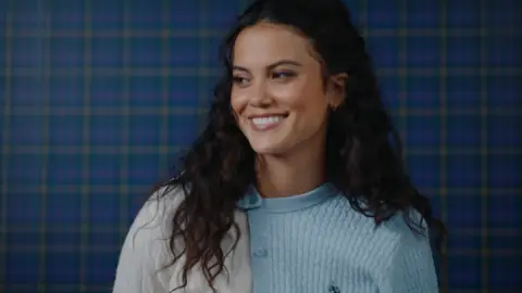 A woman with dark curly hair smiles in pale blue and white knit layers against a softly lit blue checked wall.