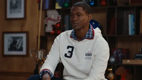 A man in a white pullover with a navy number 3 and plaid shirt collar sits at the edge of a brown leather sofa.