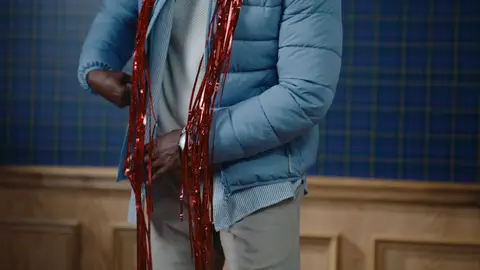 Reflective red tinsel hangs over a pale blue quilted puffer jacket and white knit top, with one hand tucked into a jacket pocket.