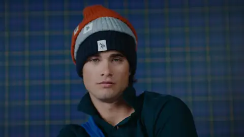 A man in a dark green top faces the camera wearing orange, gray and navy knit beanies stacked together.