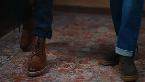 Brown leather lace-up boots and dark olive casual shoes appear below navy trousers and cuffed blue jeans on a patterned rug.