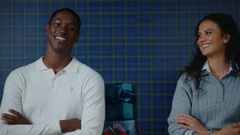 A man in a white long-sleeve polo and a woman in a gray knit top smile beside a green gift tied with a navy ribbon.