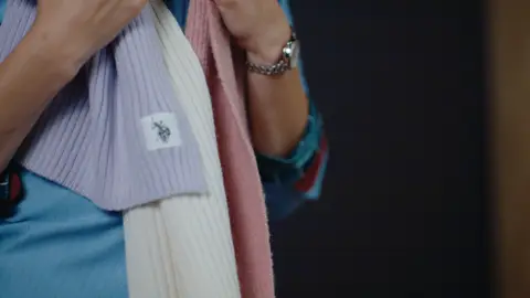 Hands hold lavender, cream and pink ribbed scarves over a blue shirt, showing a small rider-logo label and a silver wristwatch.