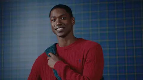 A smiling man in a red cable-knit sweater holds a green sweater draped over one shoulder against a blue checked wall.