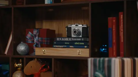 A silver and black camera rests on stacked books inside a wooden shelf, beside a small wrapped gift and silver ornament.