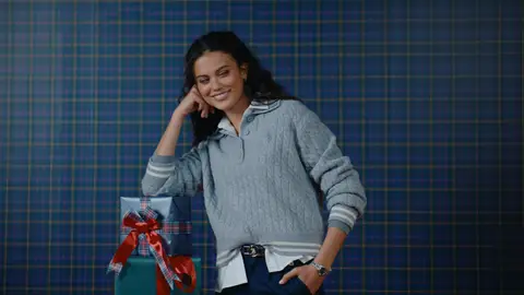 A smiling woman in a gray cable-knit top and navy trousers rests her elbow on stacked holiday gifts wrapped in plaid and solid colors.