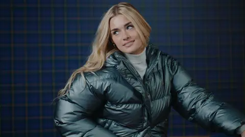 A blonde woman sits in a metallic green puffer jacket over a white turtleneck, looking to the side against a blue checked wall.