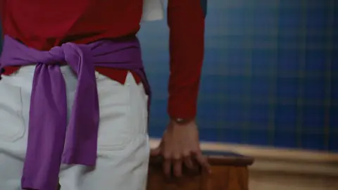 A purple sweater is tied over white trousers beneath a red long-sleeve top, with one hand resting on a wooden rail.