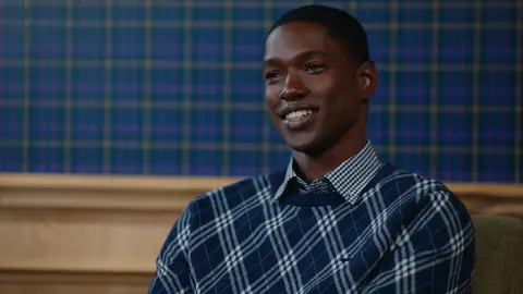 A smiling man sits in a wooden armchair wearing a navy sweater with a white diagonal plaid pattern over a small-check shirt.