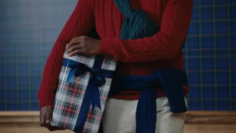 A man holds a plaid-wrapped gift tied with a navy ribbon against a red cable-knit sweater and draped green knitwear.