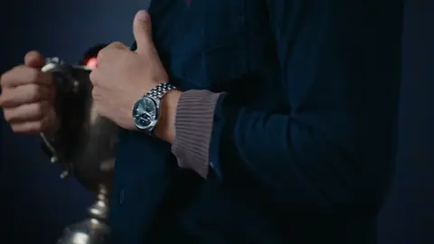 A silver bracelet wristwatch with a dark dial sits above a taupe knit cuff and navy sleeve, beside a hand holding a silver trophy cup.