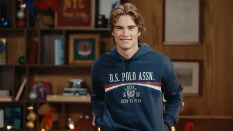 A man wears a navy graphic hoodie beside a brown leather sofa in a warmly lit room decorated for the holidays.
