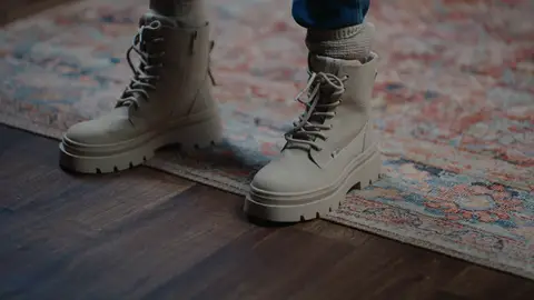 Beige lace-up boots with chunky soles, gray socks and blue jeans stand where a patterned rug meets a dark wooden floor.