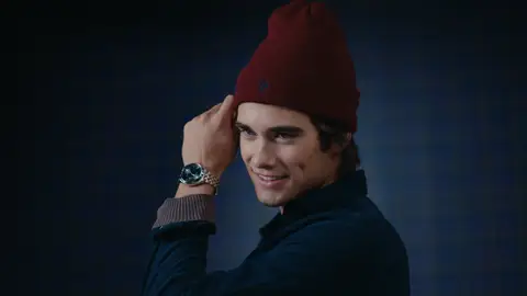 A smiling man in a burgundy knit beanie and navy shirt raises his wrist beside his face, showing a silver watch above a taupe cuff.