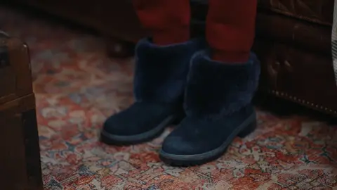 Navy fuzzy boots beneath red lounge trousers rest on a patterned rug beside a brown leather sofa.