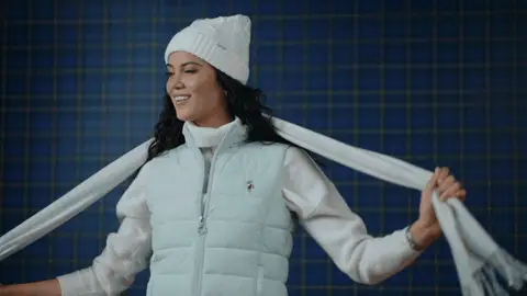 A smiling woman in a white beanie and mint quilted vest swings the ends of a pale scarf across her shoulders.