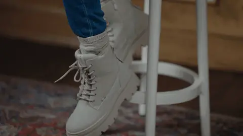 A beige lace-up boot gently swings beside a white stool above a patterned rug, with blue jeans and a gray sock visible.