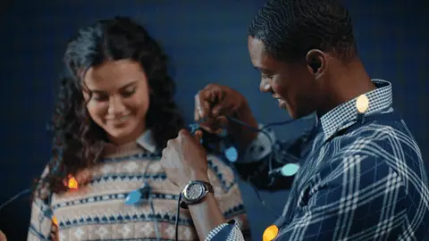 A man in a navy plaid-knit sweater raises a strand of colored holiday lights as a woman in a cream patterned sweater smiles beside him.